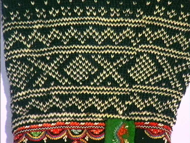 Strikket ullgarn, heimespunnet, svart og hvitt. Broderi i ullgarn. Langs nedrekanten en 13 cm bred hvit bord, resten mønsterstrikket: en bord åttebladroser, en sikksakkbord, en bord kors og kantstilte kvadrat. Langs åpningen foran og halsringingen er påsatt et stykke svart klede, saum i rosa, blått, gult og grønt zephyrgarn. Rundt ermet et 9,3 cm bredt belegg av svart klede, kantet med grønn klede 2,5 cm bred. En brodert bord langs indre og ytre kant av belegget. Aagot Noss 1969. 
