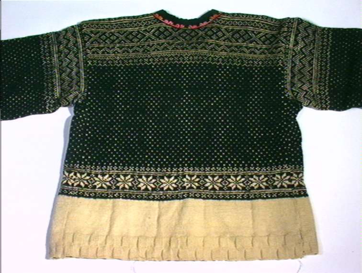 Strikket ullgarn, heimespunnet, svart og hvitt. Broderi i ullgarn. Langs nedrekanten en 13 cm bred hvit bord, resten mønsterstrikket: en bord åttebladroser, en sikksakkbord, en bord kors og kantstilte kvadrat. Langs åpningen foran og halsringingen er påsatt et stykke svart klede, saum i rosa, blått, gult og grønt zephyrgarn. Rundt ermet et 9,3 cm bredt belegg av svart klede, kantet med grønn klede 2,5 cm bred. En brodert bord langs indre og ytre kant av belegget. Aagot Noss 1969. 
