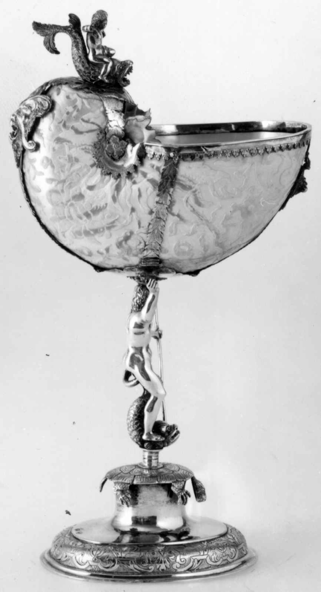 Skall av blekksprutarten Nautilus. Skåret dekorasjon, fot og innfatning sølv, støpt og gravert dekorasjon. Stpl: MF/H//1720 Morten Finchenhagen, Christiania. Skallet er brunflammet i overflaten. Nautilusblekkspruten lever i sydlige hav omkring Filippinene.