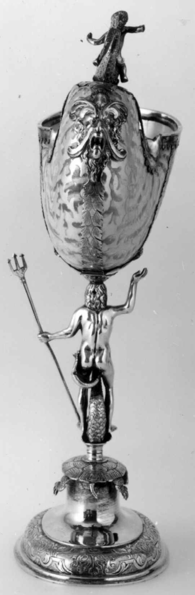 Skall av blekksprutarten Nautilus. Skåret dekorasjon, fot og innfatning sølv, støpt og gravert dekorasjon. Stpl: MF/H//1720 Morten Finchenhagen, Christiania. Skallet er brunflammet i overflaten. Nautilusblekkspruten lever i sydlige hav omkring Filippinene.