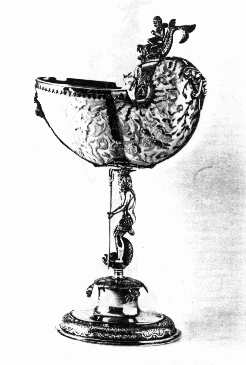 Skall av blekksprutarten Nautilus. Skåret dekorasjon, fot og innfatning sølv, støpt og gravert dekorasjon. Stpl: MF/H//1720 Morten Finchenhagen, Christiania. Skallet er brunflammet i overflaten. Nautilusblekkspruten lever i sydlige hav omkring Filippinene.