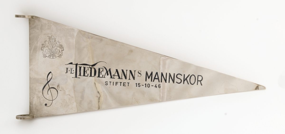 Deler til fane for Tiedemanns mannskor. Toppstykke og vimpel i metall samt transportpose i lerret.