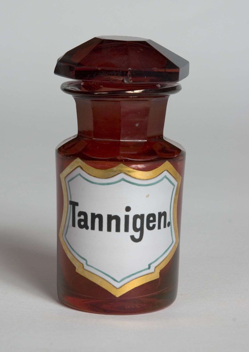 Standglass med propp. 8-kantet hals og propp. 
Hvit skjoldformet etikett med gullfarget ramme, grønn dekorlinje og svart skrift. Emaljert.