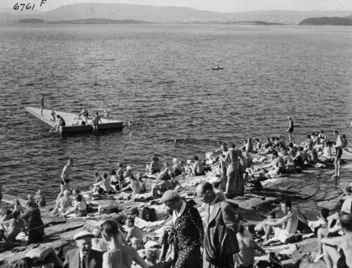Strandliv med badegjester som soler seg på svaberget ved sjøen på Ingierstrand bad utenfor Oslo. Flytebrygge i bakgrunnen.