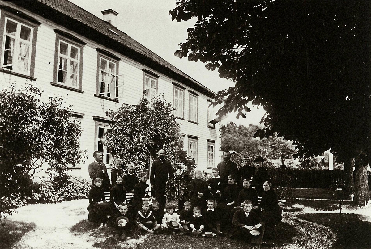 Strømsbo gård, Strømsbu, Arendal. Hovedbygningen er oppført 1760/61, men fikk sitt nåværende preg i 1804 da den ble overtatt av statshauptmann Peter Herlofson. Fredet. Fra hagesiden, hageselskap med stor familiegruppe  poserende. Navneliste, se Eksemplar 3, Påskrift.
Fra serie (NF.04551-001 til -021) fra billedmappen "Strømsbo", gård på Strømsbu i Arendal tilh. familien Herlofson. Bildene er fra ulike eiendommer og steder i Arendal tilknyttet familien, fotografert av P.A. Thorèn, Chra.