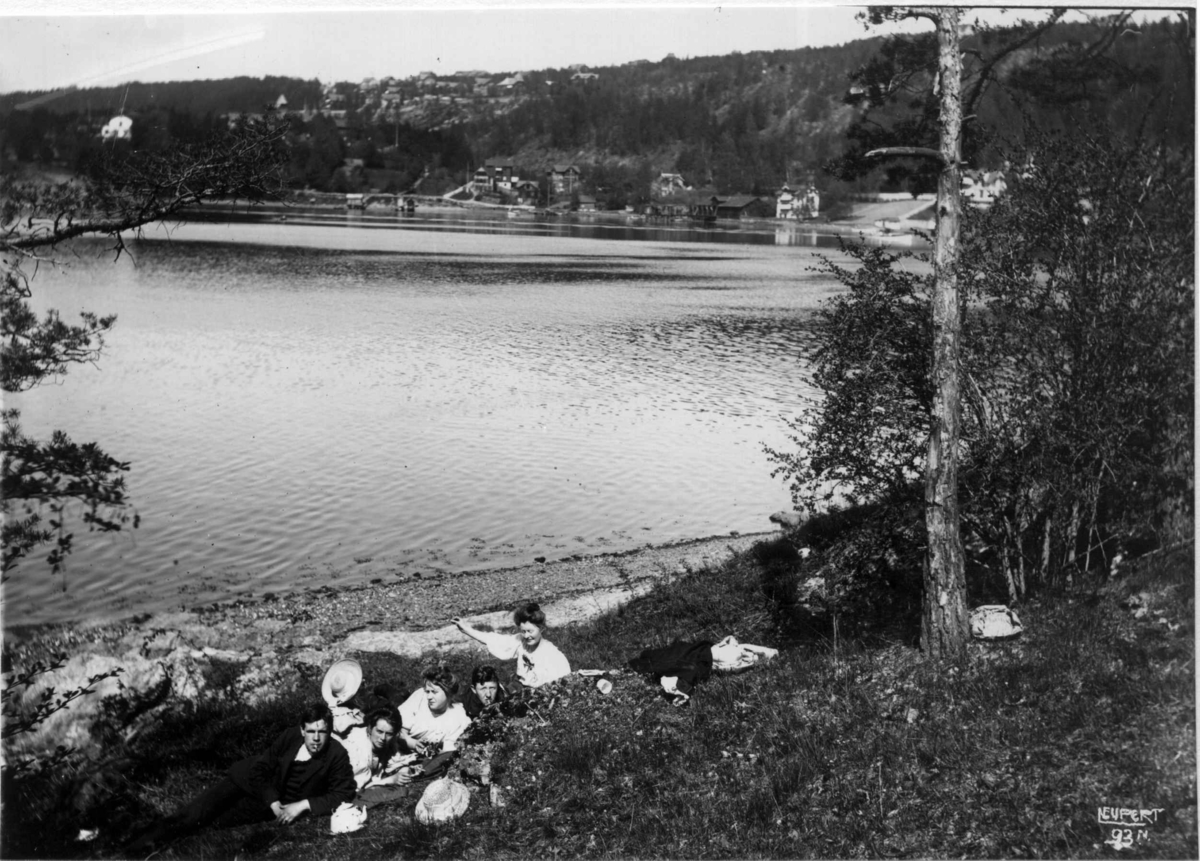 Ulvøya, Oslo 1908. Oversiktsbilde. Fjorden i forgrunnen. Gruppe med mennesker sitter på stranda. Bildet tatt mot fastlandet. Bebyggelse i bakgrunnen. 