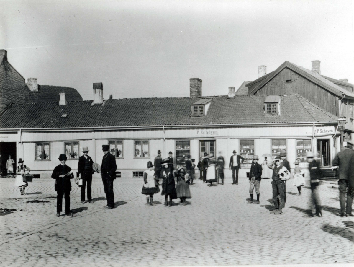 Lilletorget, Oslo ant.1880-årene. Storgårdsundersøkelser ved dr. Engelstad sommeren 1954.Mennesker på plass med brostein, foran lavt murhus med forretninger. P Schøyen, brennevinshandel.
Fra dr. Eivind S. Engelstads storgårdsundersøkelser 1954.