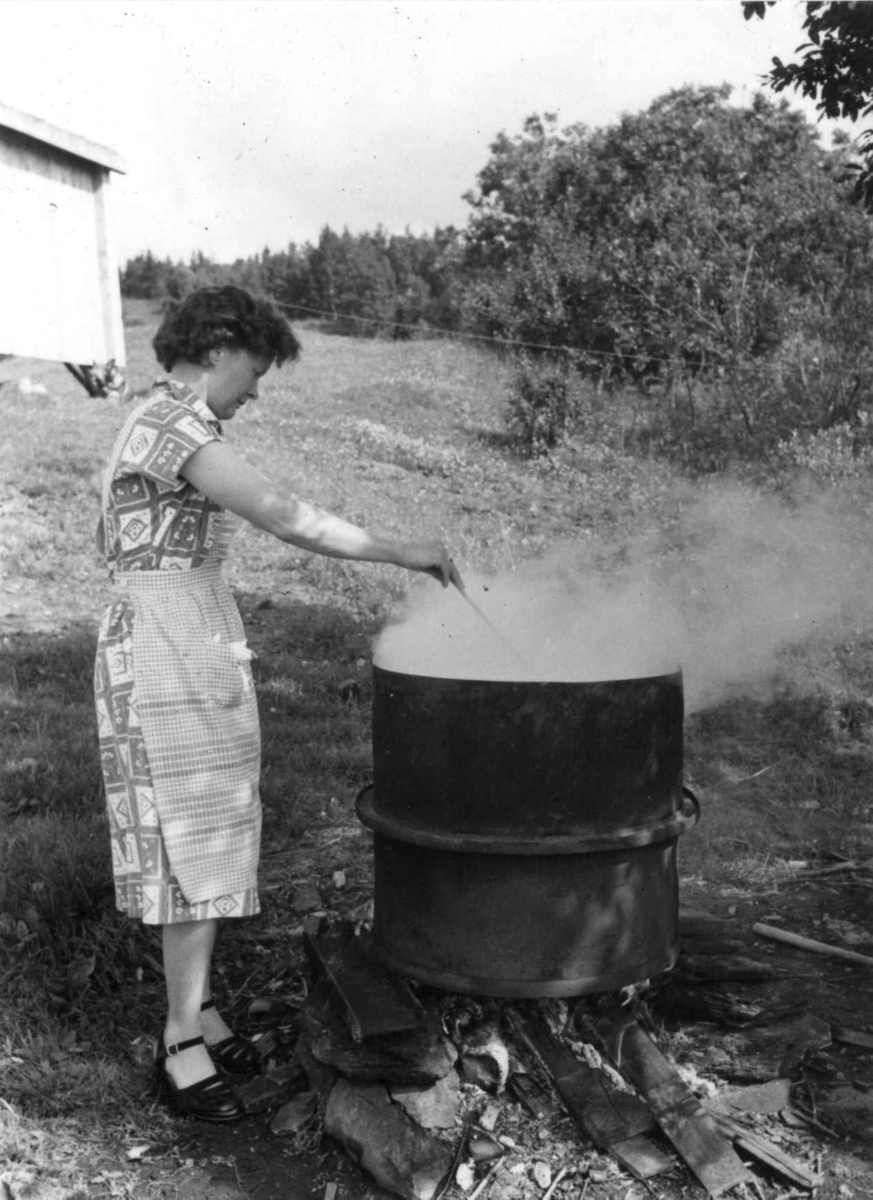 Laila Jåma vasker tøy i en jerntønne over åpen ild utendørs, Nyvikmoen 1956.