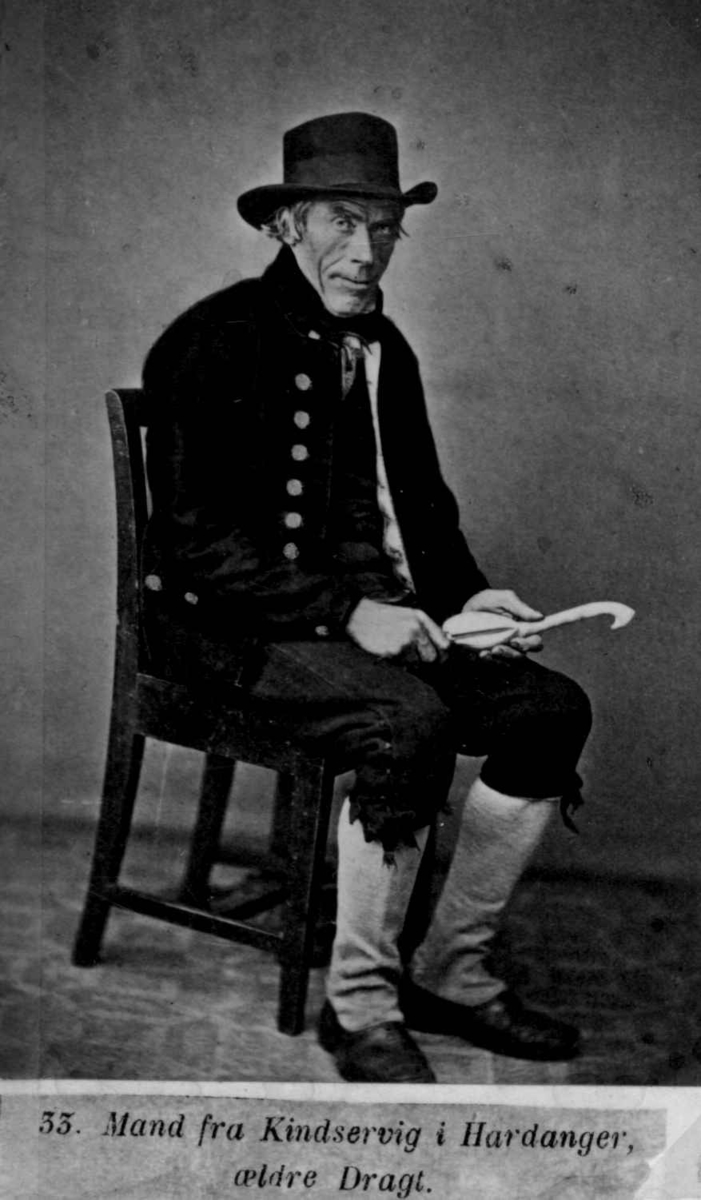 33. Mannsdrakt, Kinsarvik, Ullensvang Portrett av mann med hatt (treskjærer?) sittende i fotoatelier med nøytral bakgrunn og mønstret gulvteppe.
Fra serien "Norske Nationaldragter" (nr.33), fotografert av fotograf Marcus Selmer (1819-1900), Bergen.