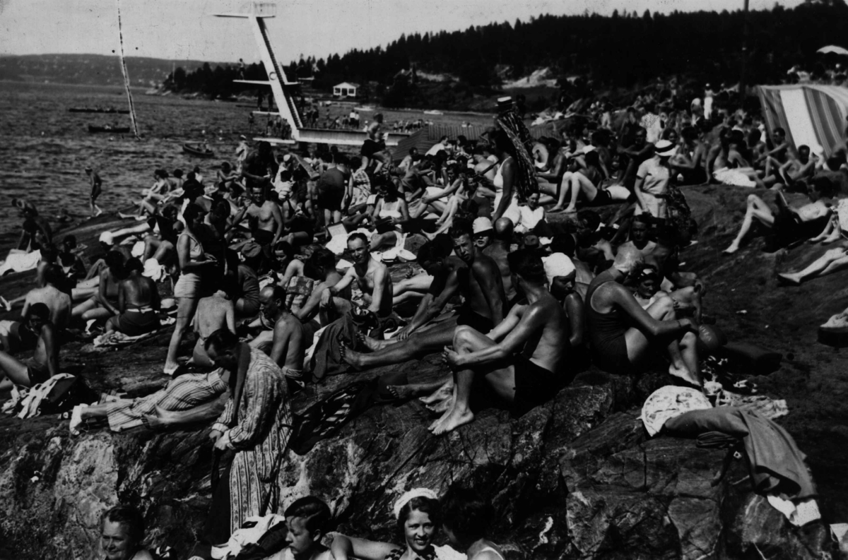 Strandliv med badegjester som soler seg på svaberget ved sjøen på Ingierstrand bad utenfor Oslo. Stupetårn i bakgrunnen.