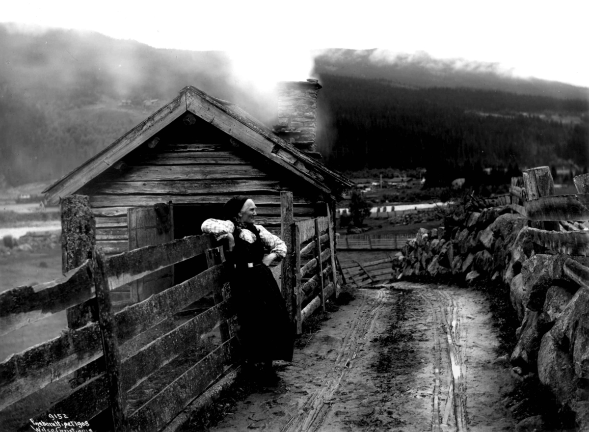 Kvinne i drakt fra Hallingdal, lent opp mot et gjerde. Hus i bakgrunnen, vei i forgrunnen.