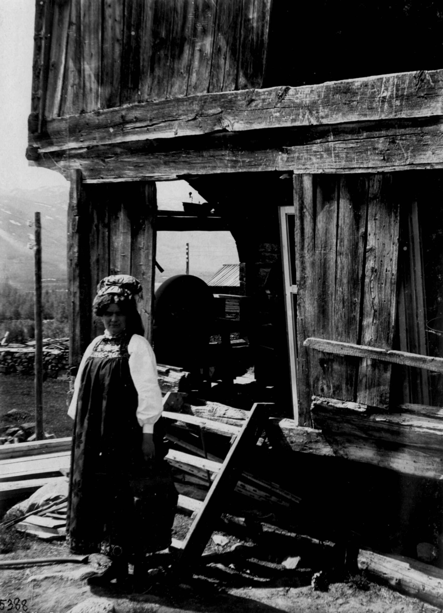 Kvinnedrakt fra Øvre Hallingdal, Buskerud. 1927. Kvinne stående foran tømmerhus.