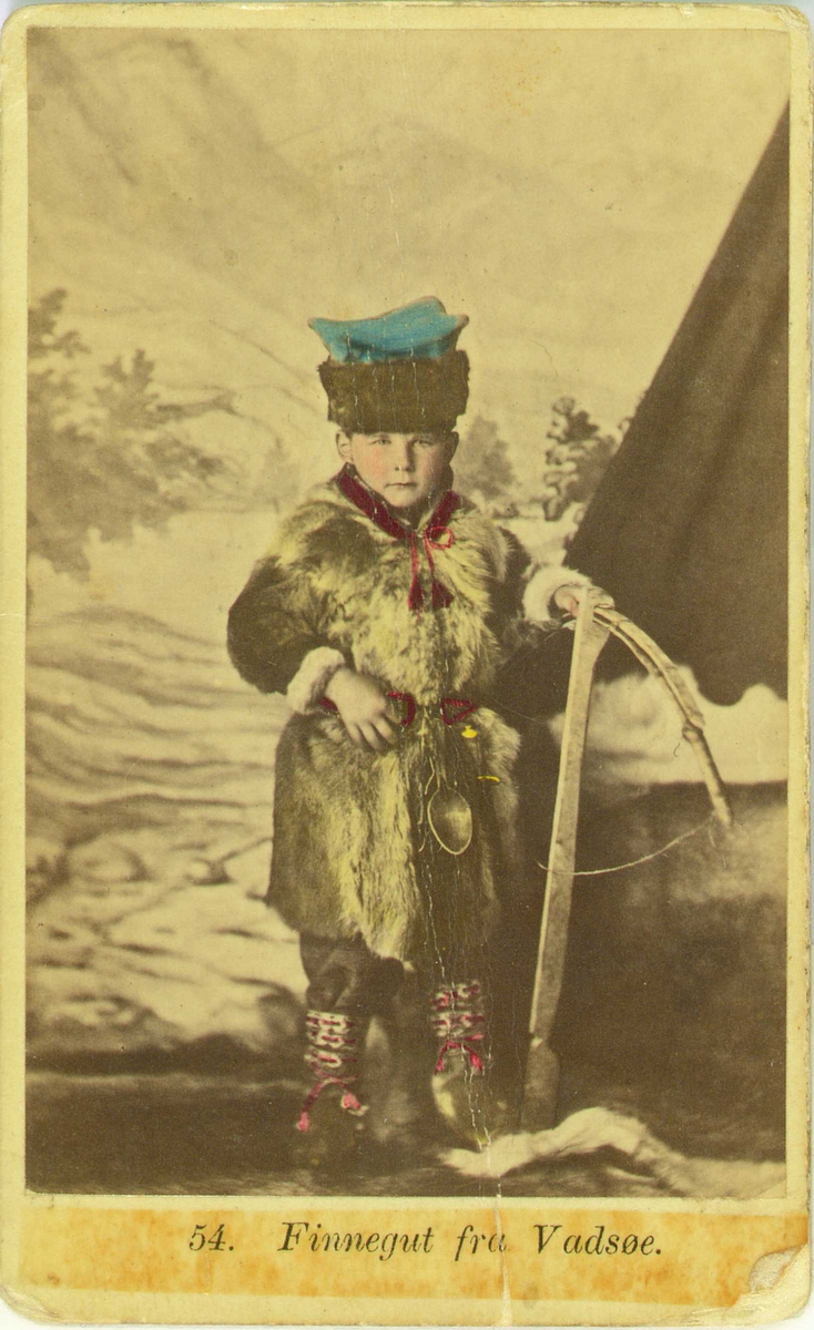 54. Guttedrakt, samedrakt, Vadsø. Portrett av gutt med pelsvest og hodeplagg ant. i fotoatelier med malt naturbakgrunn med telt (?). Gutten holder et våpen, ant. en armbrøst. Kolorert original.
Fra seien "Norske Nationaldragter" (nr.54) tatt av fotografen Marcus Selmer (1819-1900), Bergen.