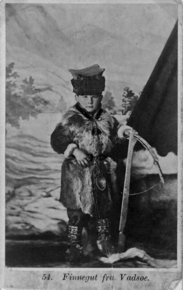 54. Guttedrakt, samedrakt, Vadsø. Portrett av gutt med pelsvest og hodeplagg ant. i fotoatelier med malt naturbakgrunn med telt (?). Gutten holder et våpen, ant. en armbrøst. Kolorert original.
Fra seien "Norske Nationaldragter" (nr.54) tatt av fotografen Marcus Selmer (1819-1900), Bergen.