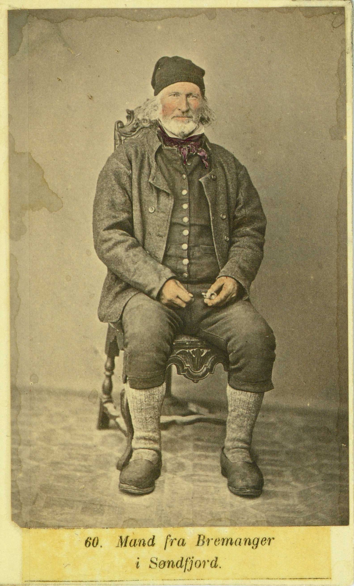 60. Mannsdrakt, Bremanger. Portrett av eldre mann sittende på stol i fotoatelier med nøytral bakgrunn og mønstret gulvteppe. Visittkortformat. Håndkolorert.
Fra M. Selmers (1819-1900) folkedraktserie "Norske Nationaldragter" (nr.60).
