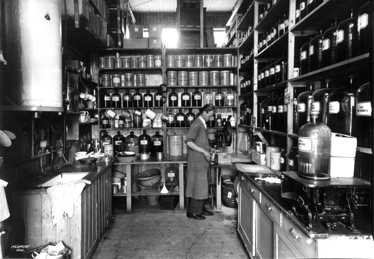 Rådhusapoteket, Oslo. 1935-1937. Provisorisk brakke. Laboratoriet. En mann i arbeid. Hyller med flasker og krukker med ingredienser.