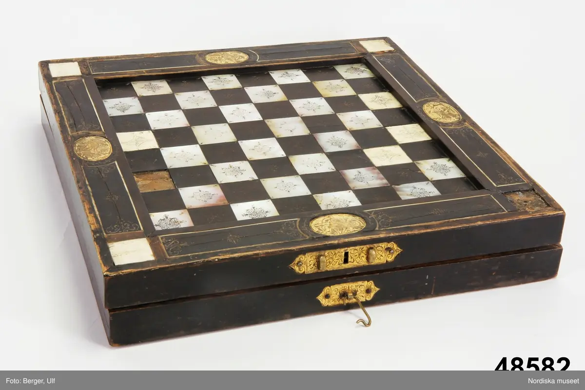Brädspelslåda för bräde, kvarn och schack, med sju spelbrickor. Svart fanér med inläggningar av pärlemor, elfenben samt graverade förgyllda metallskivor. Blindträ av ek. (Uppgifter från katalogkort). Tillhörande träask för förvaring av spelpjäserna.