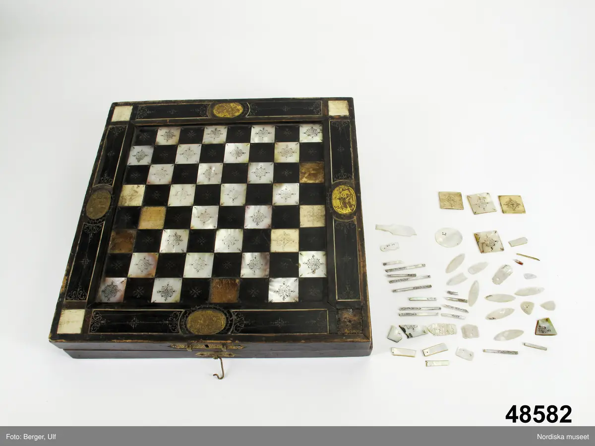 Brädspelslåda för bräde, kvarn och schack, med sju spelbrickor. Svart fanér med inläggningar av pärlemor, elfenben samt graverade förgyllda metallskivor. Blindträ av ek. (Uppgifter från katalogkort). Tillhörande träask för förvaring av spelpjäserna.