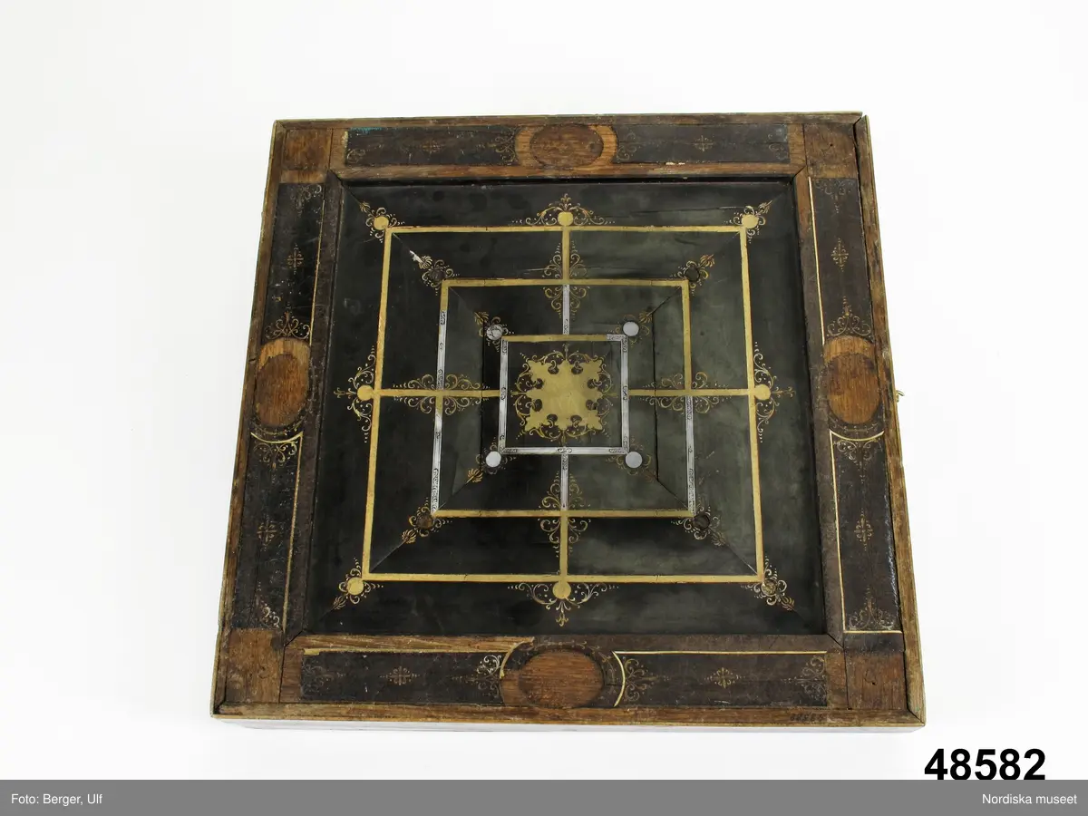 Brädspelslåda för bräde, kvarn och schack, med sju spelbrickor. Svart fanér med inläggningar av pärlemor, elfenben samt graverade förgyllda metallskivor. Blindträ av ek. (Uppgifter från katalogkort). Tillhörande träask för förvaring av spelpjäserna.