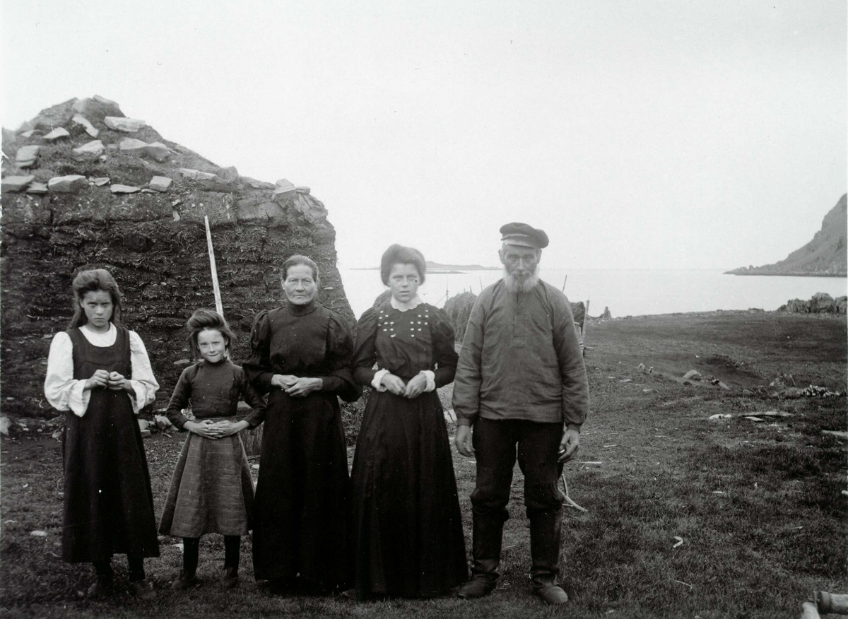 Fiskerfamilie (5) utenfor gamme, Kongsfjord, Berlevåg. Hesjer med høy i bakgrunnen. Tønne og slipestein plassert nær dem.
Del av serie fra en forskningsreise i Øst-Finnmark 1909.