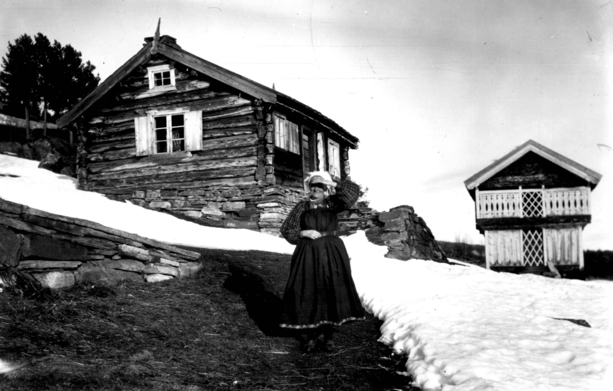 Kvinnedrakt, Ål, Hallingdal. Kvinne ses foran et
tømmerhus. Loft i bakgrunnen.