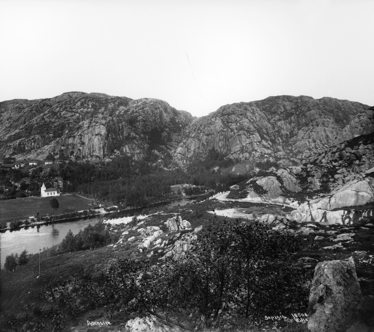 Fjellandskap, med kirke og bebyggelse ved fjordarm, Åna-Sira, Sokndal, Rogaland. Veiparti i forgrunnen.