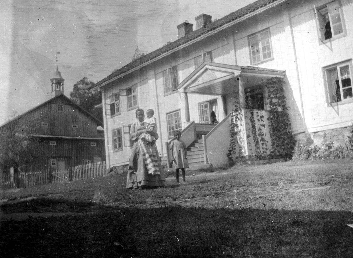 Fra gårdstun i Hurum foran våningshus i 2 etasjer med inngangsparti midt på. Bak t.v. stort uthus med klokketårn. Kvinne med to barn i forgrunnen (ukjente).
Fra malerinne Juliane Fredrikke Langbergs (1856-1930) samling, tatt ca.1895-1910 med motivkrets slekt og venner samt steder tilknyttet samvær med disse.