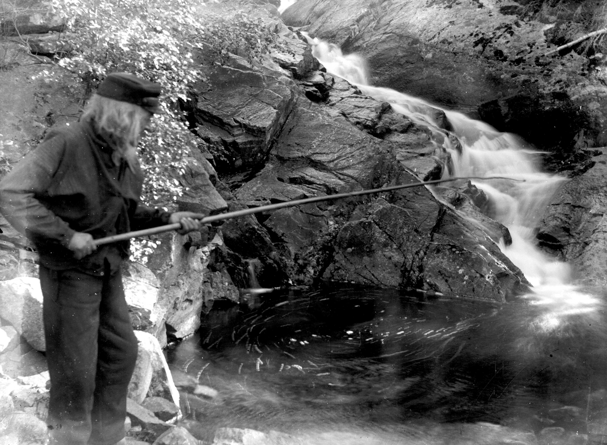 Eldre mann i skyggelue fisker (?) med en lang stang nedenfor lite fossefall.  Muligens i Eggedal.
Fra malerinne Juliane Fredrikke Langbergs samling, tatt 1895-1910 med motivkrets slekt og venner samt steder tilknyttet samvær med disse.  