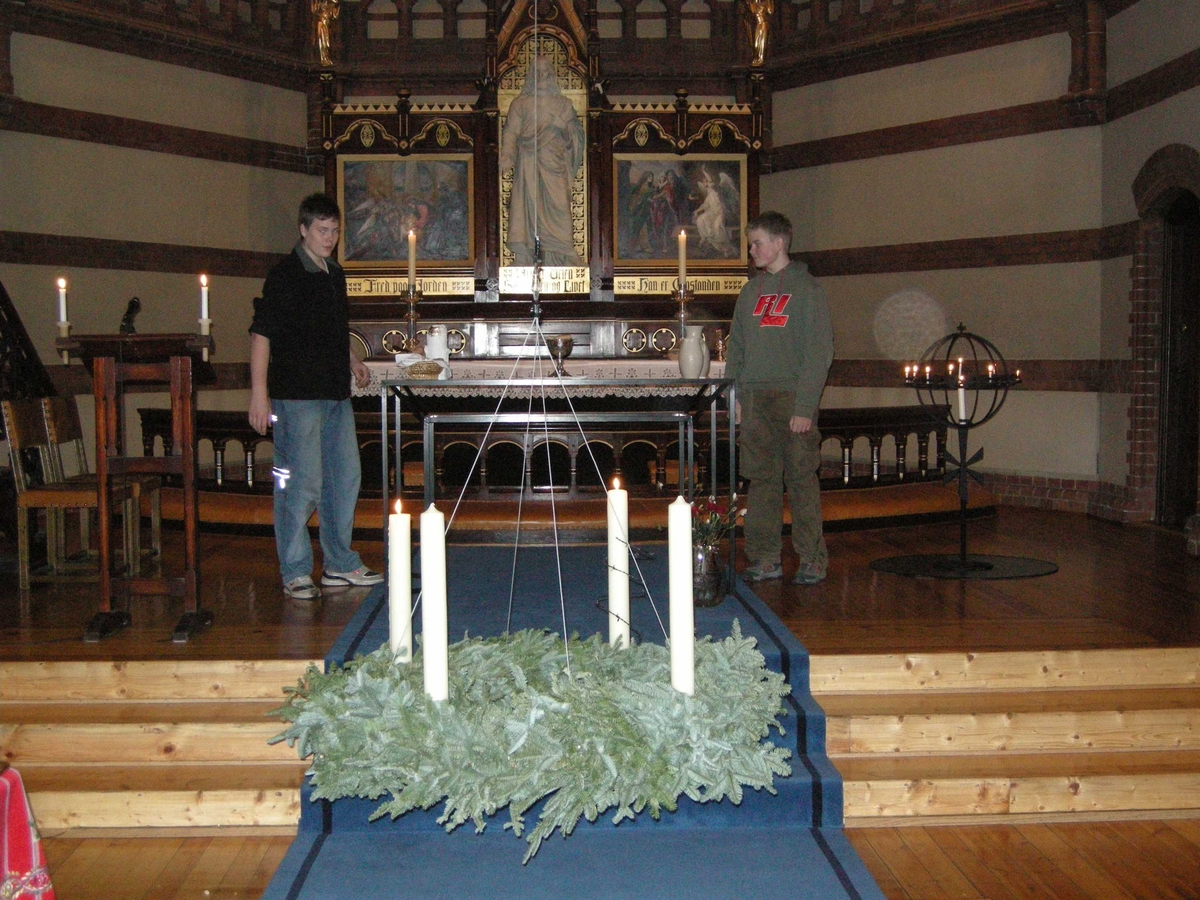 Paulus kirke i Oslo, 2. søndag i advent, 09.12.2007. To lys i lyskransen som er senket ned fra taket foran alteret er tent før den heises opp igjen. Ett lys er viklet i piggtråd. Det tennes første gang på menneskerettighetssøndagen. To konfirmanter står i bakgrunnen. Lesepult, alter, altertavle, lysglobe.