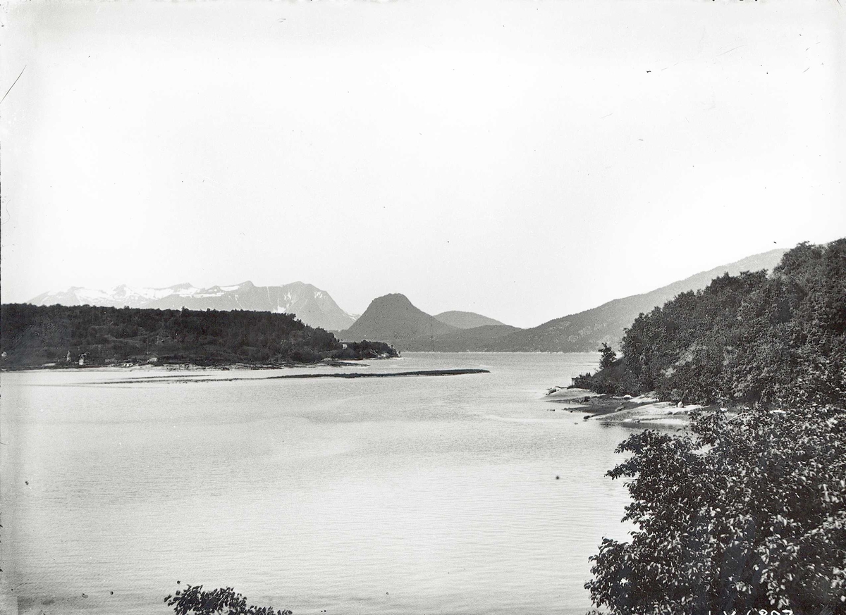 Landskap med vann, skog og snøtoppede fjell.