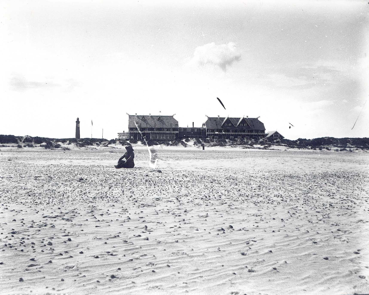 Strandhotel med dragehoder. Kvinne sitter på stor sandstrand. Fyrlykt i bakgrunnen.