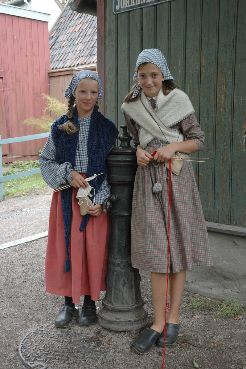 Levendegjøring på museum.
Ferieskolen uke 32 i 2005. To jenter i drakt med hvert sitt strikketøy ved vannposten på Enerhaugen på Norsk Folkemuseum.