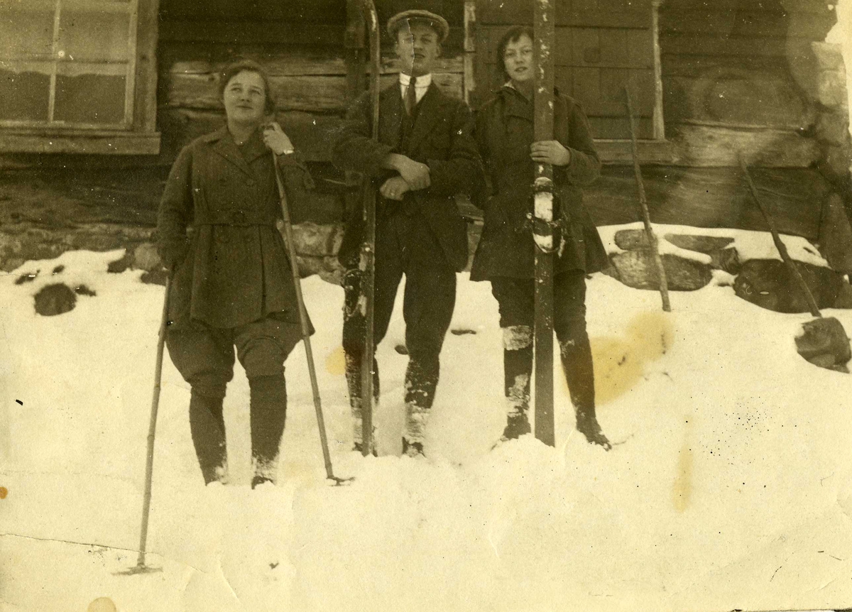 Tre ungdommer med ski foran husvegg, ca. 1917