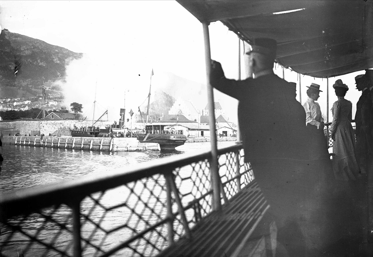 Fra dekket på en båt, mennesker ved relingen. En dampbåt ligger ved kaia. D/S Storfjord av Aalesund. Muligens Bergen havn, Rozenkrantztårnet i bakgrunnen? 1902.