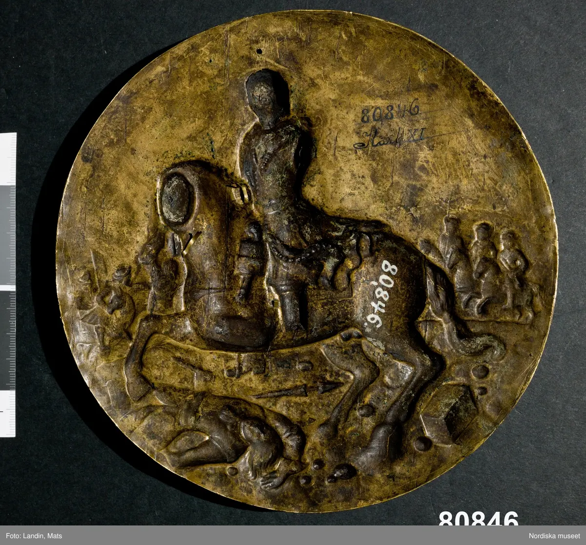 Katalogkort:
"Platta, porträttmedaljong, mässing, förgylld. 1676 [med blyerts]. Drivet arbete, hög relief. Carl XI till häst med dragen värja å slagfältet. Till höger å en fyrkantig sten läses: 'Carolus XI R. S. Praeli Lond. [Cond?] 4 dec. 1676.' I rund förgylld träram med pärlkrans. Uttagen ur ramen."
KHq [=Kersti Holmquist]