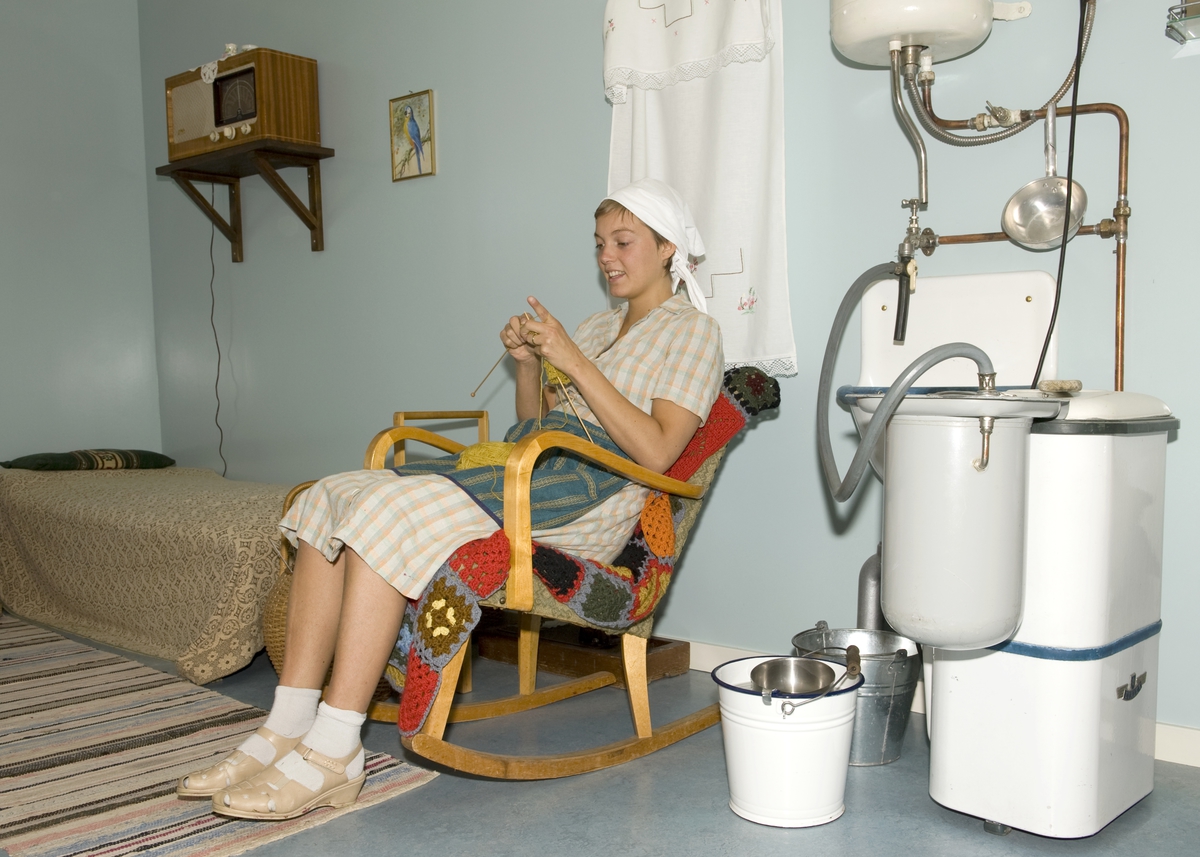 Tunvert som strikker i hovedbygning fra Stiklestad, Trøndelagstunet på Norsk Folkemuseum, august 2010. Interiør med radio og vaskemaskin. Formidling av boskikk og dagligliv i et 1950-talls gårdsbruk i Trøndelag. 