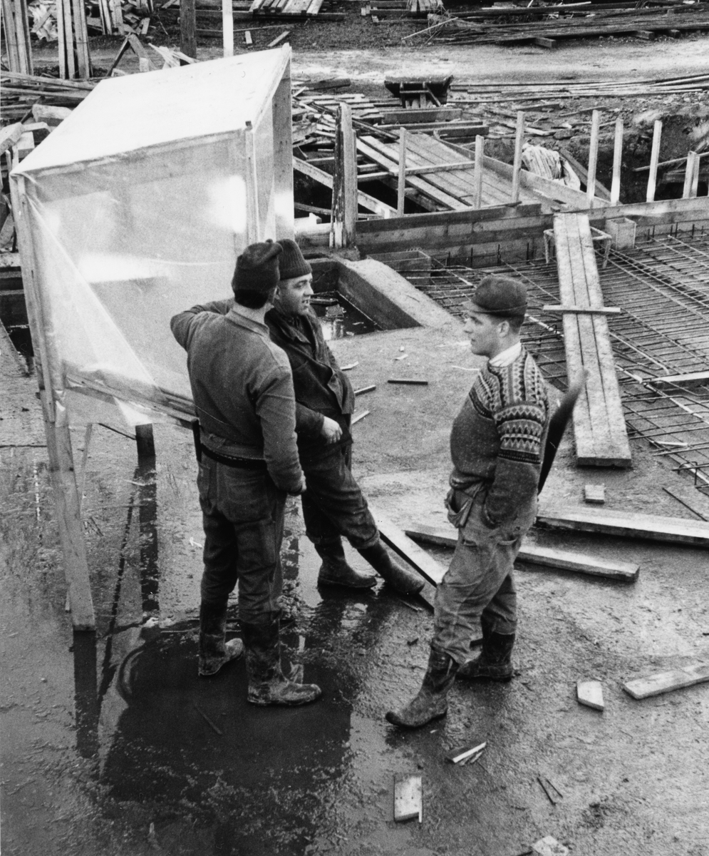 Byggeplass.
Konstruksjon av Tiedemanns Tobaksfabrik på Hovin i 1967. Tre stk bygningsarbeidere står på byggeplassen å snakker.