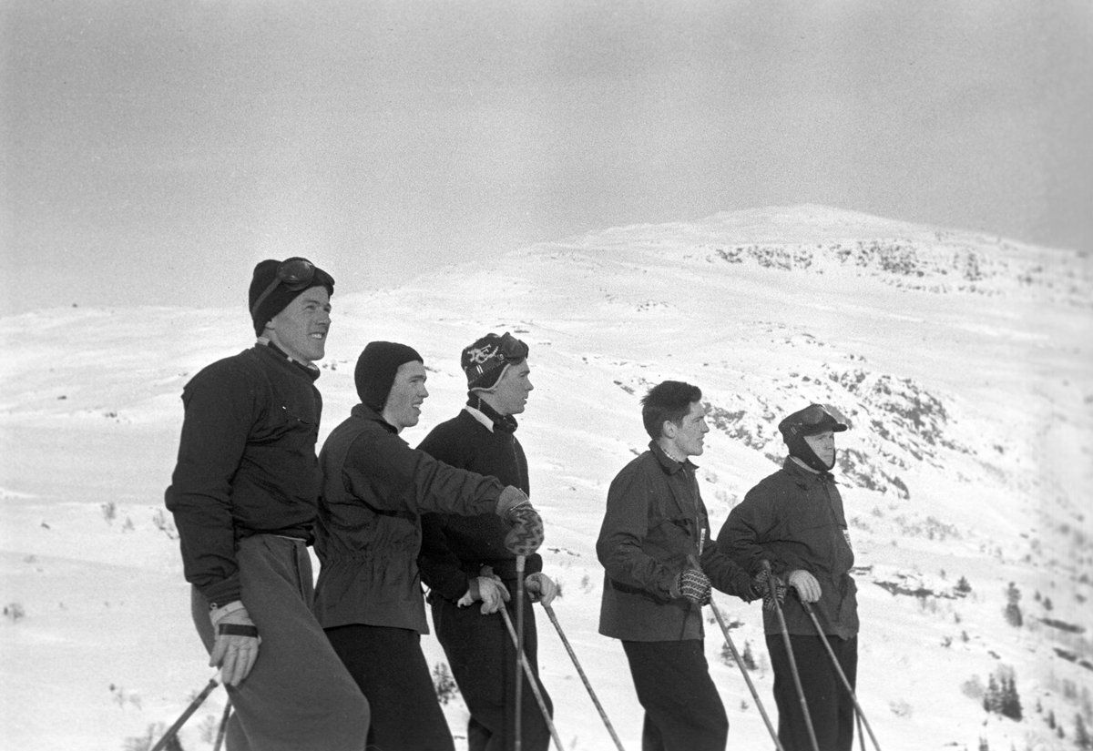 Fem personer med snødekte fjell i bakgrunnen. Holmenkollrennene på Voss 1952. Fotograf Dagbladet