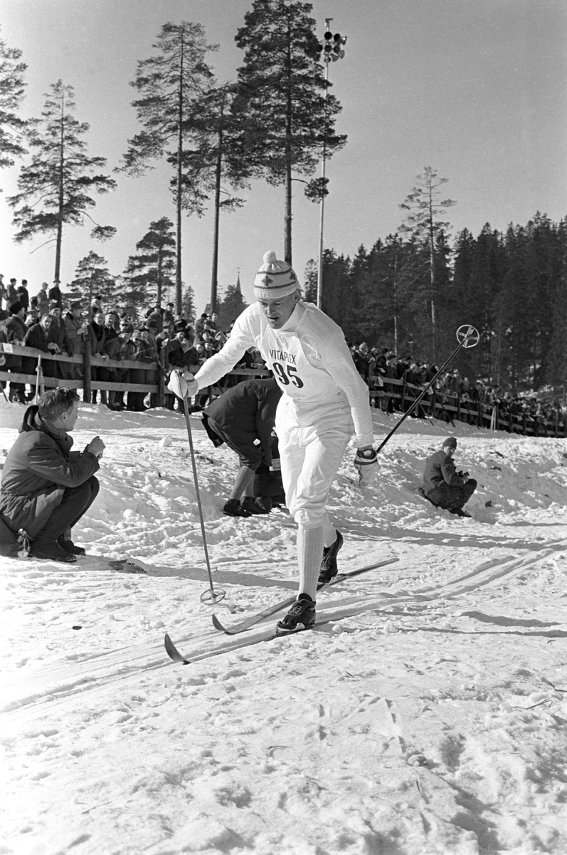 Skiløper med startnummer 95 i Holmenkollen. Publikum langs løypa.  Pressefotografer på sidelinjen. Holmenkollrennene 1963.