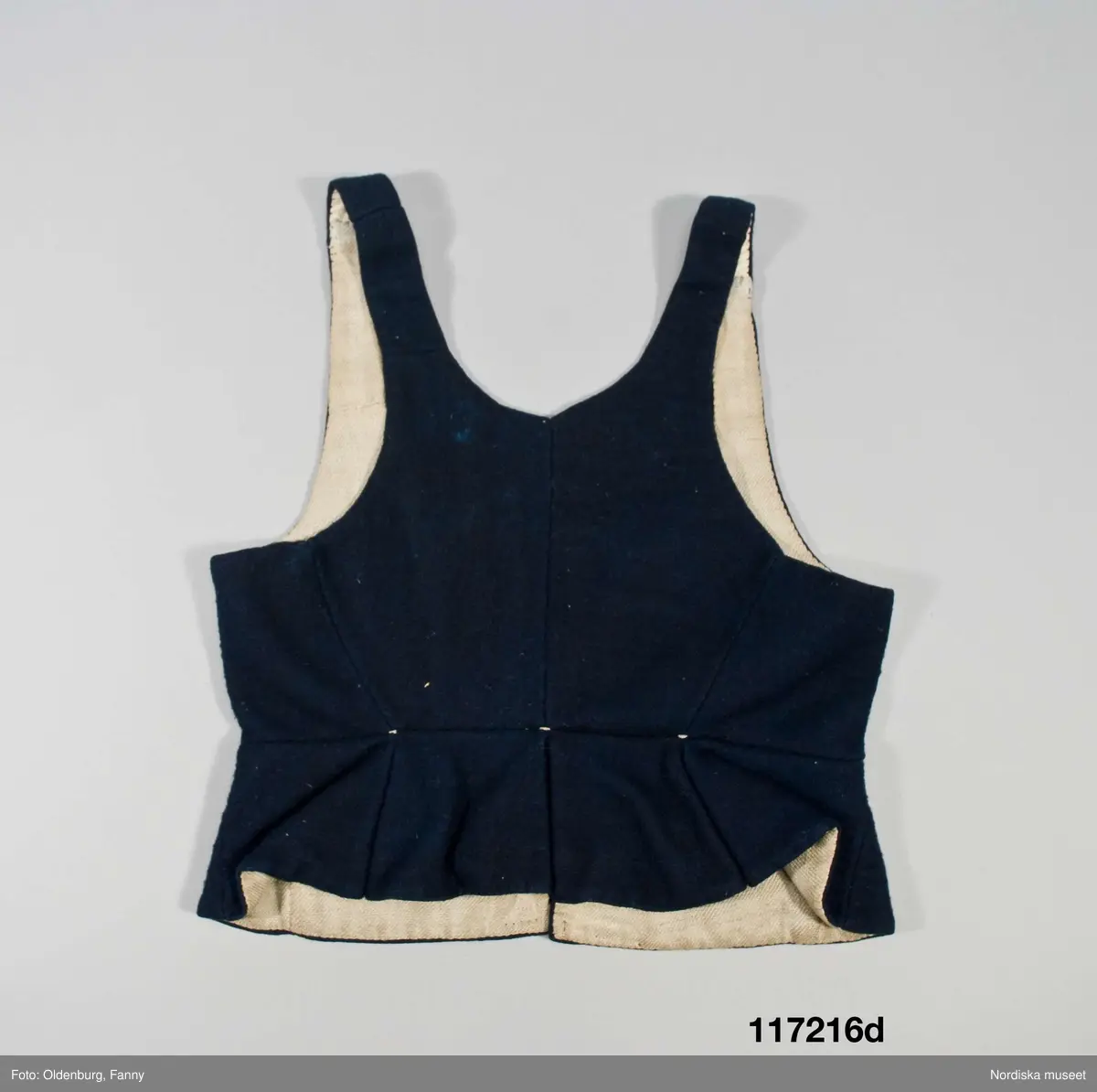 Huvudliggaren:
"Sorgdräkt för kvinna. Särk med 'hängseltåg', 'särkyvdel'; vit -'häsklä'; blått livstycke; svart tröja; huvudduk; 'svart näsduk'; hatt; 'raskförklä', 'sockar',  'hårbandsvifsa', huvudbindel."

Bilaga:
a: "8 Särk, övdel. av linne eller blågarn. med broderade linningar på överdelen"
c: "11. Hvit häsklä."
Anm: Kung Kerstin Larsdotters resa i Floda sn Dalarna 24/2 - 8/3 1912. Innehåller förteckning på inköp av dräktdelar från Floda, inv nr 117 216 - 117 239. Här finns  tillverkningstid och beskrivningar på vissa av dräktdelarna.  I vissa fall nämns också utförligt på vilka helgdagar dräktdelen  användes.

Katalogkort:
a: Hängselsärk
b. Överdel av halvlinne vävt i kypert.
c.  Halskäde av småmönstrat bomullstyg, liknande kambris, vävt  i kypertvariation med drällmönster.
d. Livstycke av mörkblå kraftig vadmal,
e. Tröja av grov svart vadmal

i. 
 Mönstervävt band med upplockat mönster i rött z-tvinnat ullgarn på vit linnebotten, med hjärtan, kryss och rombformer, 2 band om vardera 124 cm har vikts och sytts ihop så att det ena bildar en ögla och det andra har fria ändar avslutade med en tofs i bandets garn.
Läs mer om dräktskicket i Floda i: Albert Alm, Dräktalmanacka för Floda socken, Nordiska museet, Sthlm 1930
/Berit Eldvik 2005-04-14