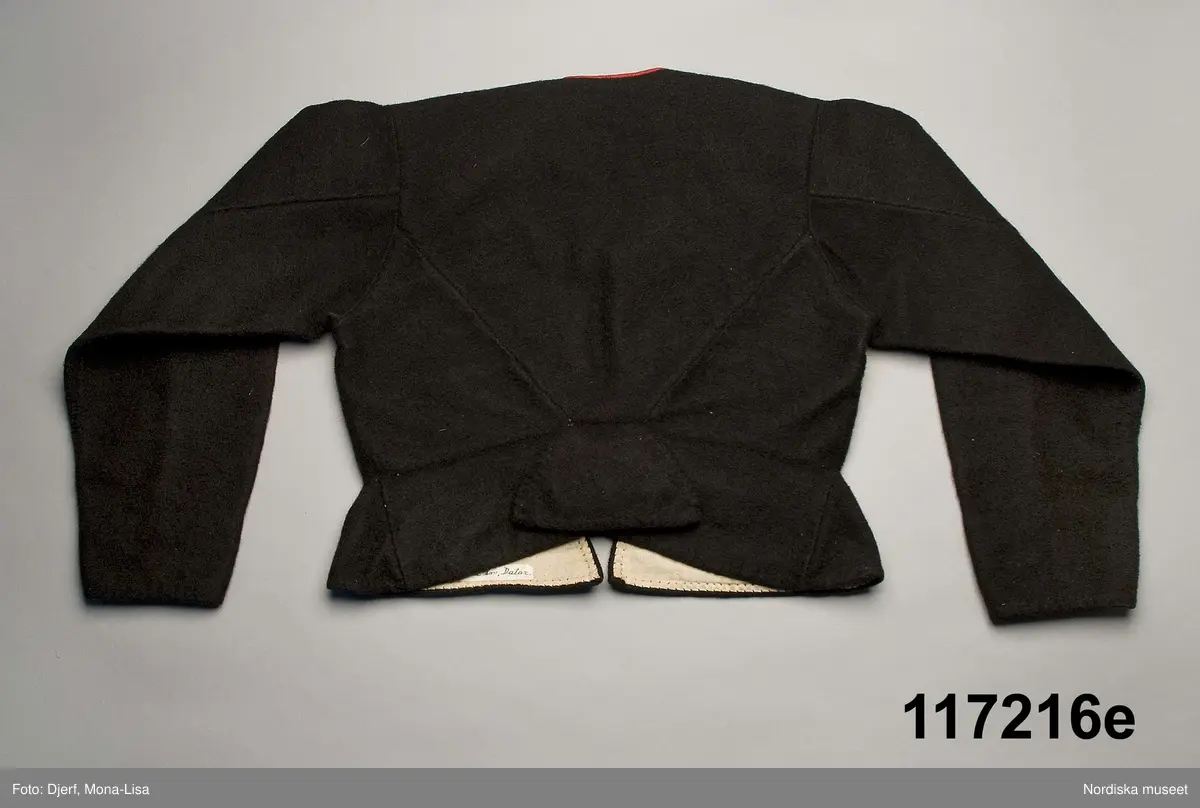 Huvudliggaren:
"Sorgdräkt för kvinna. Särk med 'hängseltåg', 'särkyvdel'; vit -'häsklä'; blått livstycke; svart tröja; huvudduk; 'svart näsduk'; hatt; 'raskförklä', 'sockar',  'hårbandsvifsa', huvudbindel."

Bilaga:
a: "8 Särk, övdel. av linne eller blågarn. med broderade linningar på överdelen"
c: "11. Hvit häsklä."
Anm: Kung Kerstin Larsdotters resa i Floda sn Dalarna 24/2 - 8/3 1912. Innehåller förteckning på inköp av dräktdelar från Floda, inv nr 117 216 - 117 239. Här finns  tillverkningstid och beskrivningar på vissa av dräktdelarna.  I vissa fall nämns också utförligt på vilka helgdagar dräktdelen  användes.

Katalogkort:
a: Hängselsärk
b. Överdel av halvlinne vävt i kypert.
c.  Halskäde av småmönstrat bomullstyg, liknande kambris, vävt  i kypertvariation med drällmönster.
d. Livstycke av mörkblå kraftig vadmal,
e. Tröja av grov svart vadmal

i. 
 Mönstervävt band med upplockat mönster i rött z-tvinnat ullgarn på vit linnebotten, med hjärtan, kryss och rombformer, 2 band om vardera 124 cm har vikts och sytts ihop så att det ena bildar en ögla och det andra har fria ändar avslutade med en tofs i bandets garn.
Läs mer om dräktskicket i Floda i: Albert Alm, Dräktalmanacka för Floda socken, Nordiska museet, Sthlm 1930
/Berit Eldvik 2005-04-14