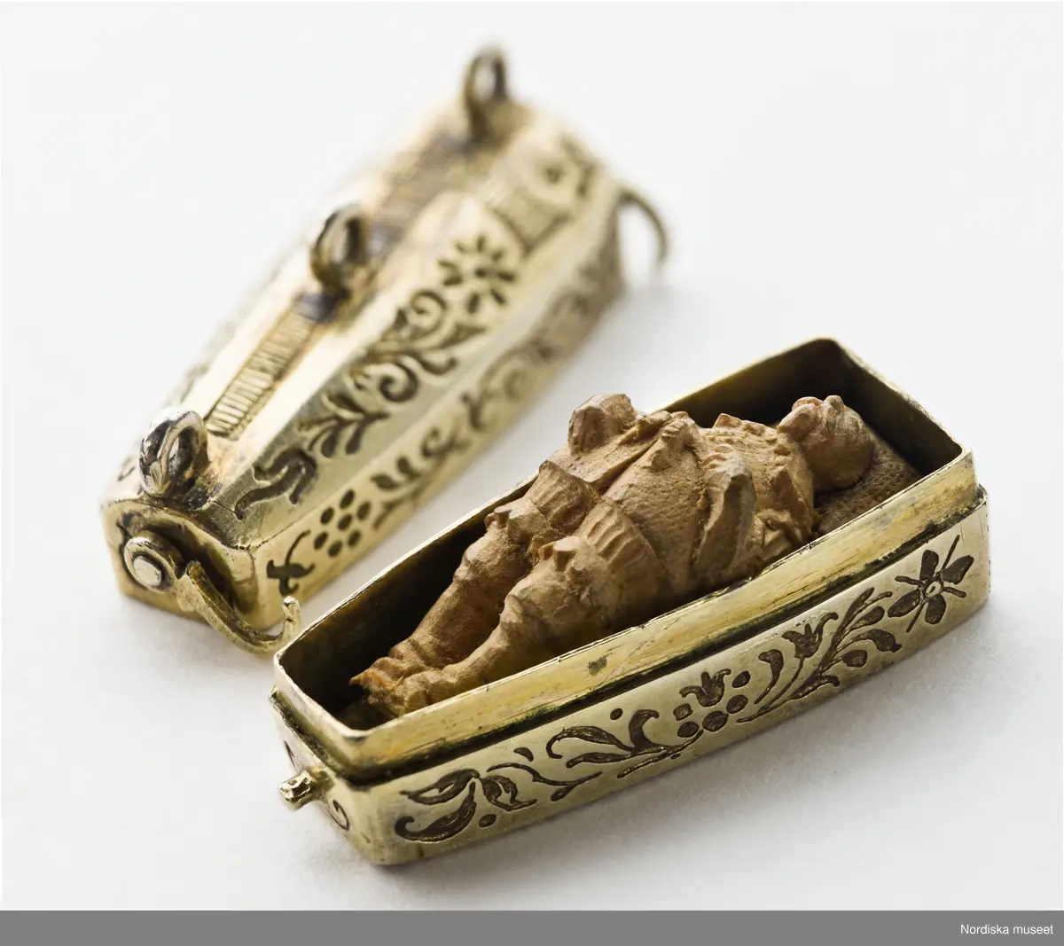 Katalogkort:
"Smycke (berlock)
Silver
L 3,5 cm, br. 1,2 cm
I form av Gustav II Adolfs likkista. Trycklock med haspar. Graverad dekor. Miniatyrbild i trä av kunge, fäst i botten."