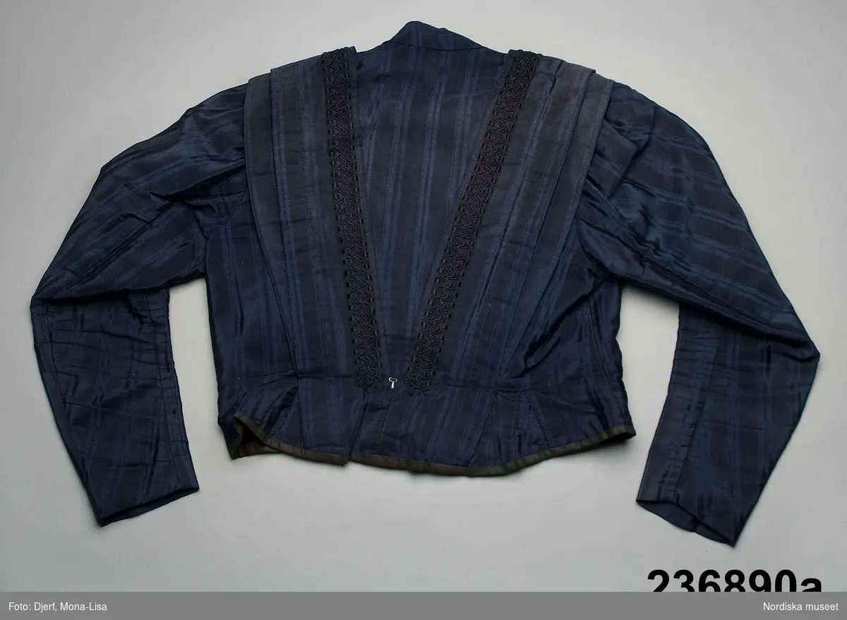 2-delad klänning av mörkblått randvävt halvsiden. a-b samt löst skärp c.

a. Liv figursytt på foderstomme av  ljusgul bomullskypert i fiskbensmönster. 2 framstycken och ett ryggstycke med ett rundat skört bak som dock stoppas ned i kjolen, Fram djupt V-ringad med  2 breda lagda veck som  löper i V-form från axel ned mot underkanten både fram och bak. Längs med vecken även en 3 cm bred  svart maskingjord spets . I ringningen isättning av samma tyg med en mängd tvärgående stråveck, tättslutande ståndkrage  styvad med något mellanlägg. Isydd lång ärm, svängd med 2 sömmar, något rynkad på ärmkullen, snäv vid handleden.
Mitt bak i midjan en fastsydd häkta att fästa mot kjolen. 
b. Kjol av 7 sneddade våder, sydd på foderstomme av svart bomullslärft s.k domestik. Framvåden markerad med nedsydda veck vid sömmarna. Kjolen slät framtill och rynkad bak i midjan mot en 2 cm bred linning av samma tyg. Mitt bak i linningen en fastsydd hyska att häkta mot livet. Dolt sprund i vänster framsöm knäppt med  5 tryckknapapr och överst en hake och hyska. Infälld ficka i sprundet. Skoning i nederkanten av samma tyg som fodret. I kanten slitband med plyschkant.
Anm. 10-12 cm av tyget invikt upptill vid linningen. Kanske ett sätt att inte klippa bort tyg om man skulle vilja återanvända det senare. Det var mycket vanligt att just tygrika kjolar kunde sys om till annat.
c. Löst skärp av samma tyg.  Veckat på längden. Ofodrat, söm mitt bak, 3 fastsydda stålfjädrar på insidan. Knäpps med 3 svarta hakar och tränsade trådöglor 
Klänningen bör vara sydd efter 1903 då Prym uppfann tryckknappen.
/Berit Eldvik 2011-09-08