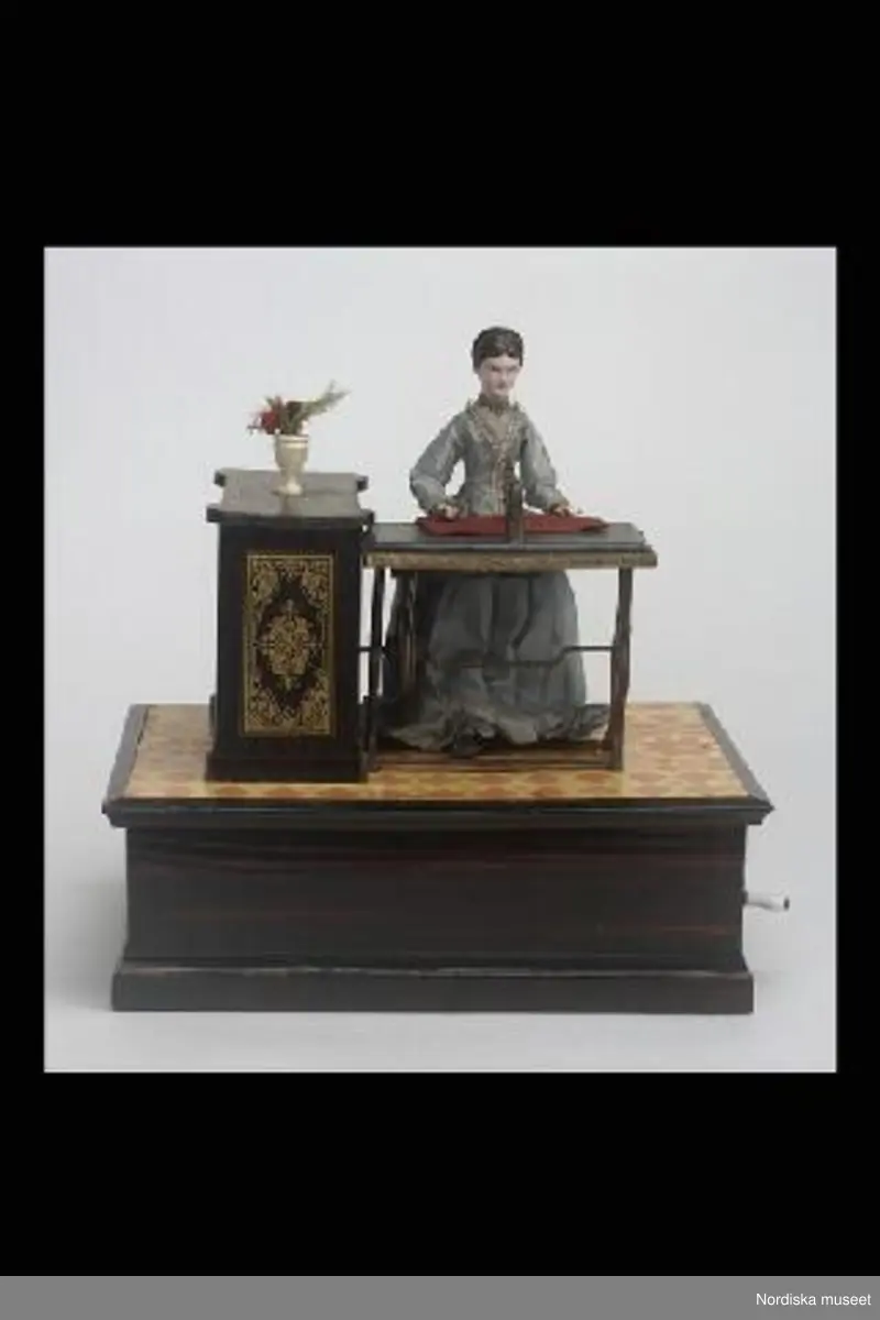 Inventering Sesam 1996-1999:
L  24,8   B  17,7   H  25 (cm)
Speldosa (a), leksak, damdocka sittande vid symaskin på rektangulär, vevf¿rsedd låda av brunbetsat och lackat barrträ.
Docka av målad papier mach¿ med brunt uppsatt hår, ljusblå sidenklänning med turnyr, trampsymaskin av brunmålat trä; gråmålat bord med konturerat underrede, trampa och vevhjul, symaskinsarm med nål, r¿tt tygstycke, träpall på tre ben med r¿d sammetssits, skåp av brunbetsat och lackat trä med dekor i guldfärgat ¿verf¿ringstryck, kruka av trä med blomma av papper, textil och fjäder, imiterad korkmatta av lackat papper med tryckt geometriskt m¿nster i gult och orange. 
F¿rvaringslåda med skjutlock (b+) av obehandlat barrträ, tryckt etikett med motiv av leksaker samt diverse påskrifter, bl a "1/12 dz 1782".
Vid gång r¿r sig fottrampan, nålen går upp och ned och tyget f¿rflyttar sig.
Ägarinnan fick speldosan i gåva av sina f¿räldrar när hon var barn på 1890-talet, då familjen bodde i Kalmar. M¿jligen tysk tillverkning.
Anna Womack feb 1998