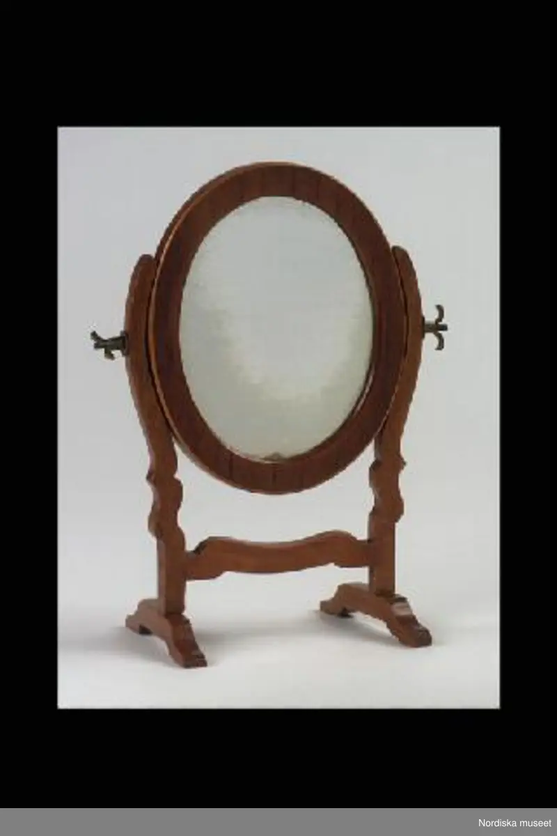 Inventering Sesam 1996-1999:
H 17 cm
B 15 cm
Oval svängbar spegel, ram trol. av mahognyfan¿r, benställning med vridbara av mahogny, spegeln är fäst mellan två ståndare med två mässingsskruvar.
Skänktes till Carolina Dorotea Öhmann (1840-1935) som flicka av fröken Gjörwell.