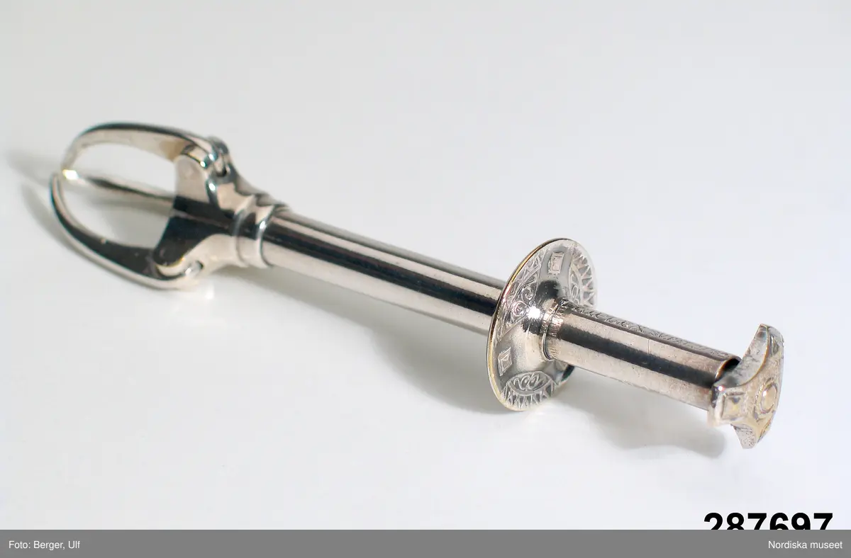 Huvudliggaren:
"Sockertång av nysilver; cylindriskt handtag, tre gripklor, som öppnas vid tryck på en trepassformad platta; stpl 'Triplex Sv. Pat. 27782. L. 10,7 cm.
Från giv. föräldrarhem. Från 1920-talet.
G. 13/4 1971 Fröken Ida Genman, Vällingby."