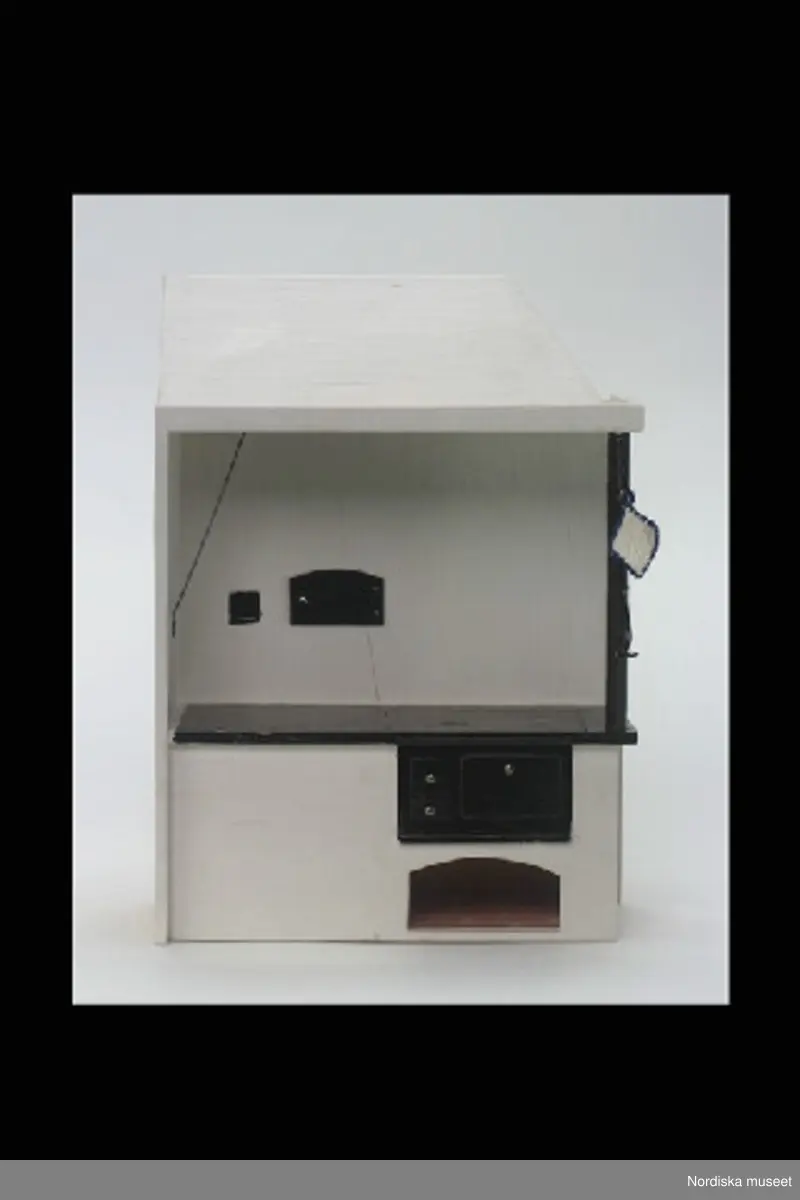 Inventering Sesam 1996-1999:
a) Grytlapp, fyrkantig, virkad av tunt vitt bomullsgarn, blå kant och hank. L 1 cm, B 1,7 cm.
b) Spiskrok, av svartmålad vitmetall, krok i ena änden, ögla i den andra. L 5,3 cm, B 0,8 cm. 
c) Spis med kupa, av trä, rektangulär form. Vitmålad spiskåpa, bakvägg och underdel. Spishäll och vedspis svartmålade.  Spjällstång av vitmetall. Under vedspisen urtag för ved, målat för att imitera tegelstenar. I urtaget ligger 11 vedträn (vedträn saknas vid inv. 1997). Spisen stämplad på baksidan "NOLBYN / BB / Värmländskt hantverk". L 15,2 cm, B 7,2 cm, H 21 cm.
d) Vedkorg med ved. Flätad av spån. I korgen 16 st vedträn. H 5 cm, B 3,8 cm. (korg och ved saknas vid inventering 1997).
Hör till dockskåp inv.nr 298.529, (köket). Se dess kat.kort för mer info.
Berith Bergström, Stockholm, ritade och tillverkade dockskåpsinventarier från 1930-tal till 1960-tal under namnet "Nolbyn". Bergström hade en affär "Värmländskt hantverk" på Sibyllegatan, Stockholm. Föremålen är tillverkade efter svenska förebilder i skala 1:10.
Dockskåpet brukat av systrarna Christina (f 1948) och Margareta (f 1949) Hofman-Bang i Djurholm, döttrar till givarinnan.
Bilaga; uppgifter om tillverkaren.
BR juni 1976 [=Berit Rönnstedt]
Leif Wallin okt 1997