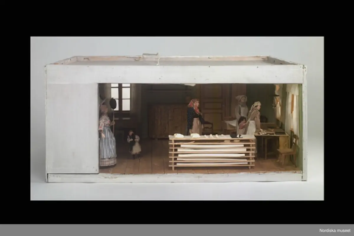 Inventering Sesam 1996-1999:
L 70 cm
B 55 cm
H 31,5 cm
Dockskåp, tittskåp förställande sekelskifteskök vid storbak/julbak, med dockor och inredning.
Rektangulär låda, vitmålad utsida tre fönster med fönsterglas av plast, en dörr. Framsidan bestående av glasskiva. Invändigt grönmålade väggar, vitt tak, skurgolv, vita gardiner av textil.. 
Till vänster vedspis (trä) med vit spiskåpa. Baktill i rummet skänk och bord, till höger slagbord med bakplåtar. Flera stolar (bl a av Lindometyp) i rummet. 6 st dockor, fyra kvinnor (av plast) i sekelskifteskläder och två små flickor (av massa, en hållande en katt). Framtill i rummet, staplade på varandra, träskivor med tyg och bröd av massa (brödet står på jäsning).
All inredning och dockor fastklistrade i golvet (ev något ändrat i museet, jmfr bilagan)
Skåpet försett med elektrisk belysning (glödlampa i framkant) och vit sladd med svart kontakt.
Tillverkat av Berith Bergström, 1896-1979 Lidingö, som under 1930-70-tal ritade och tillverkade dockskåp och dockskåpsinventarier och sålde under namnet Nolbyn i sin affär Värmländskt hantverk, Sibyllegatan i Stockhom. 
Jmfr dockskåp inv.nr 298.259 och se dess bilaga för mer info.
Bilaga. = om Berith Bergström
Leif Wallin feb 1998