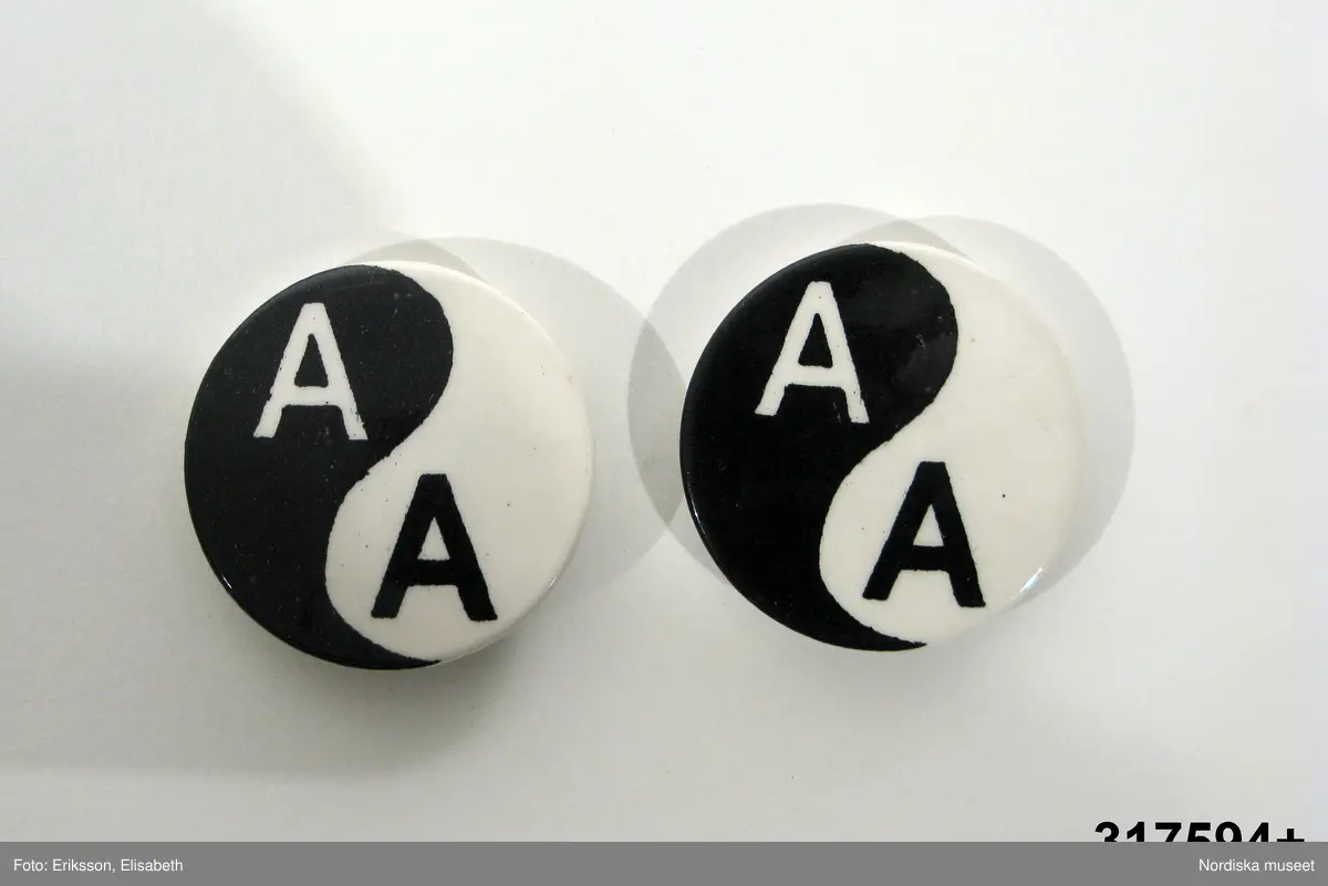Huvudliggaren:
"Rockslagsmärke, metall. Runt svart/vitt, 2 st, yin/yang märke med monogram AA (anti apartheid). Gåva av sydafrikan till givaren 1961 under vistelse i England. Nål, krylle. Använd i Stockholm.
Margit Forsberg f. 1921 bodde under en kortare tid sommaren 1961 i England i samma familj som en sydafrikan. Han berättade om förhållandena för de svarta i sitt hemland, vilket grep henne djupt. Hon fick 2 märken av honom, som sedan bars hemma i Stockholm.
/Berit Eldvik 2009-12-18