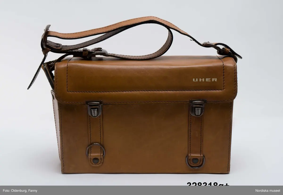 a-g:
Bärbar bandspelare för rullband. Metall, rektangulär, grå eloxerad, tangenter, vred på långsidan, fönster med ljudskala, varvräknare. Spole, mikrofon, mikrofonstativ, bruksanvisning. Batteri- och nätdriven, laddningsbar.
Tillhörande brun läderväska, axelväska med bred rem, ficka för bruksanvisning, mikrofonfack, konstmaterialfoder, uppknäppbar framsida, fällock med spännen. Adresslapp handskriven med förste intendet Göran Rosanders handstil.

a+) Bandspelare
b) Batteriladdare med sladd
c) Mikrofon med sladd
d) Mikrofonstöd
e) Intruktionsbok
f) Kopplingsschema (papper)
g+) Väska.

Den bärbara Uher-bandspelaren innebar en revolution för fältarbete. Fältarbetaren var inte enbart hänvisad till papper och penna. På 1970-talet fanns till en början endast en bärbar bandspelare. Fältarbeten planerades efter tillgänglighet till bandspelare. Samtliga avdelningar samsades till en början om dennna.
/Heidi Henriksson 2005-10-11